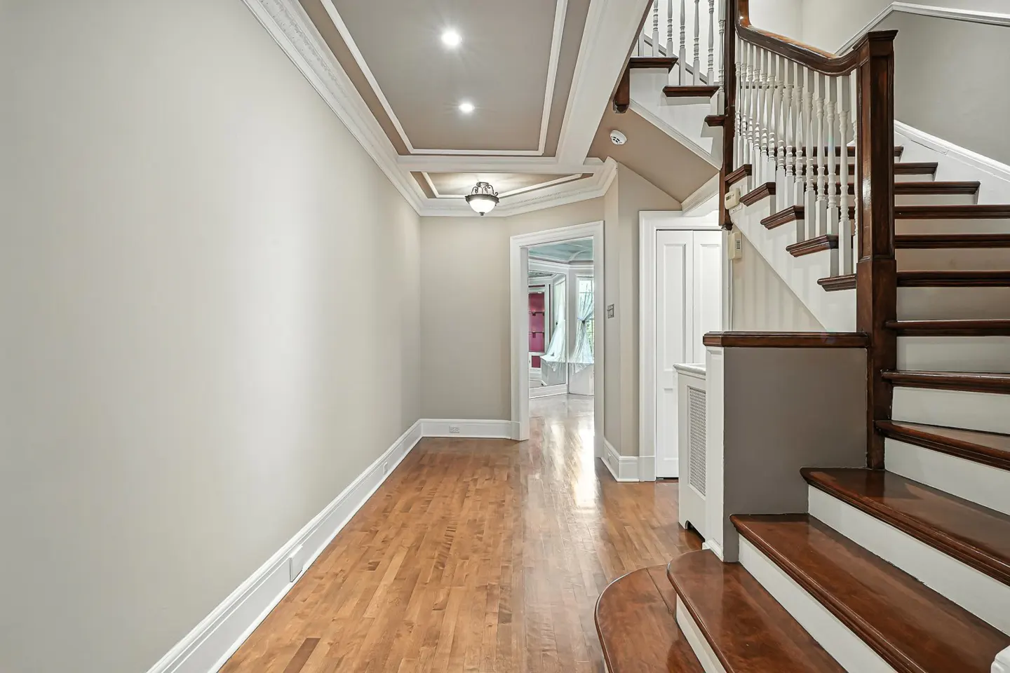 Hallway with hardwood floors, beige walls, and a staircase with dark wood railings. A doorway leads to another room.