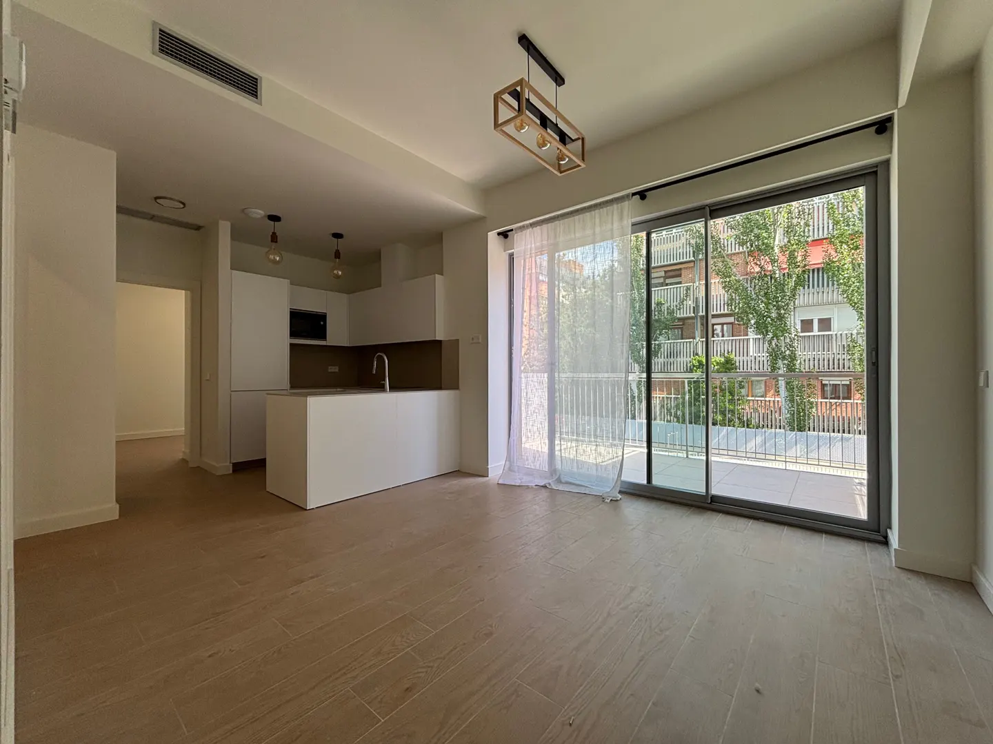 Bright, modern apartment with wood floors, white kitchen island, and sliding glass doors to a balcony overlooking a building with trees.