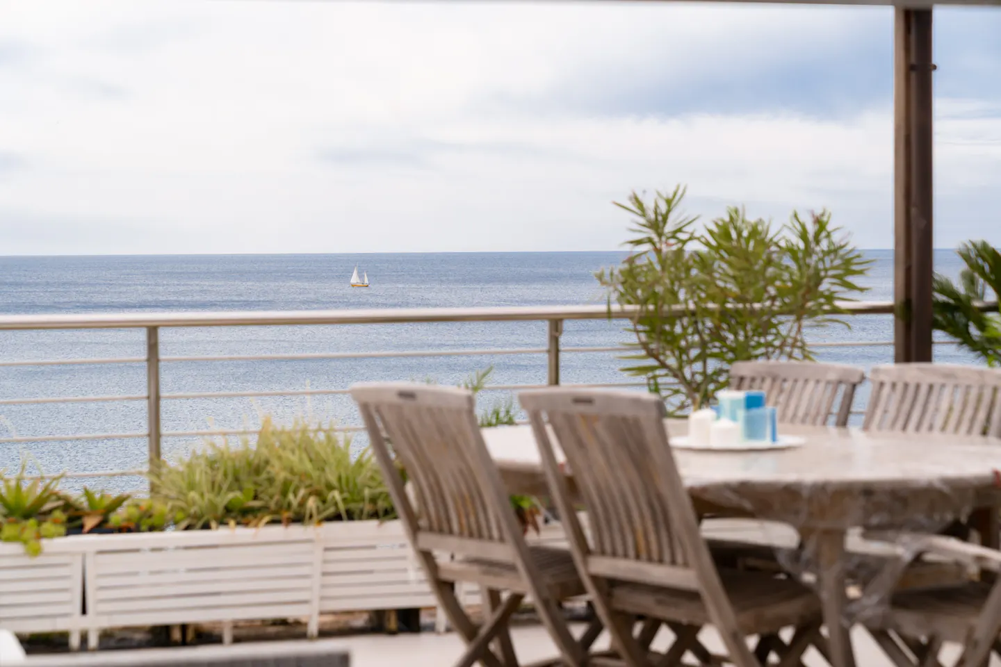 Outdoor patio with wooden table and chairs overlooking the ocean. A sailboat is visible in the distance.
