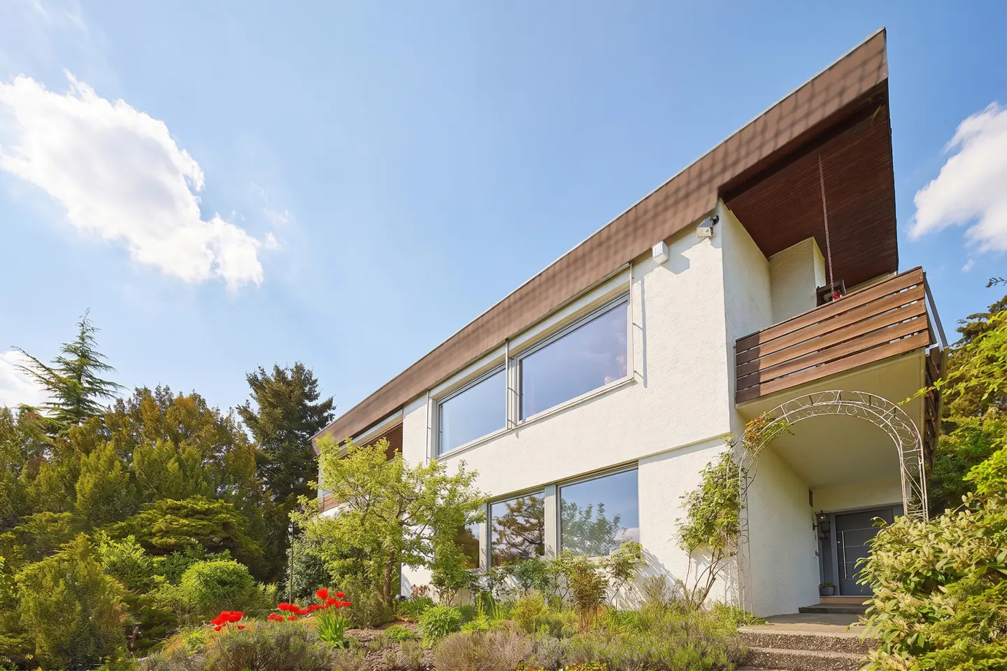 Modern two-story home with white stucco walls, large windows, a wooden balcony, and a lush garden under a blue sky.