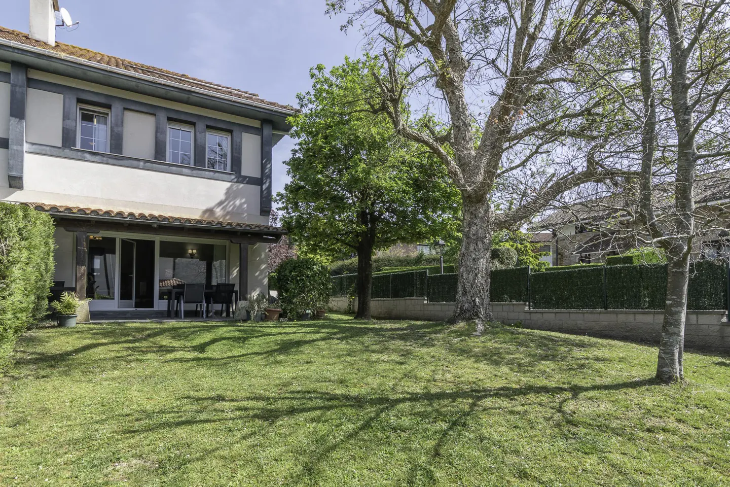 Backyard view of a two-story house with a patio, green lawn, trees, and a hedge fence.