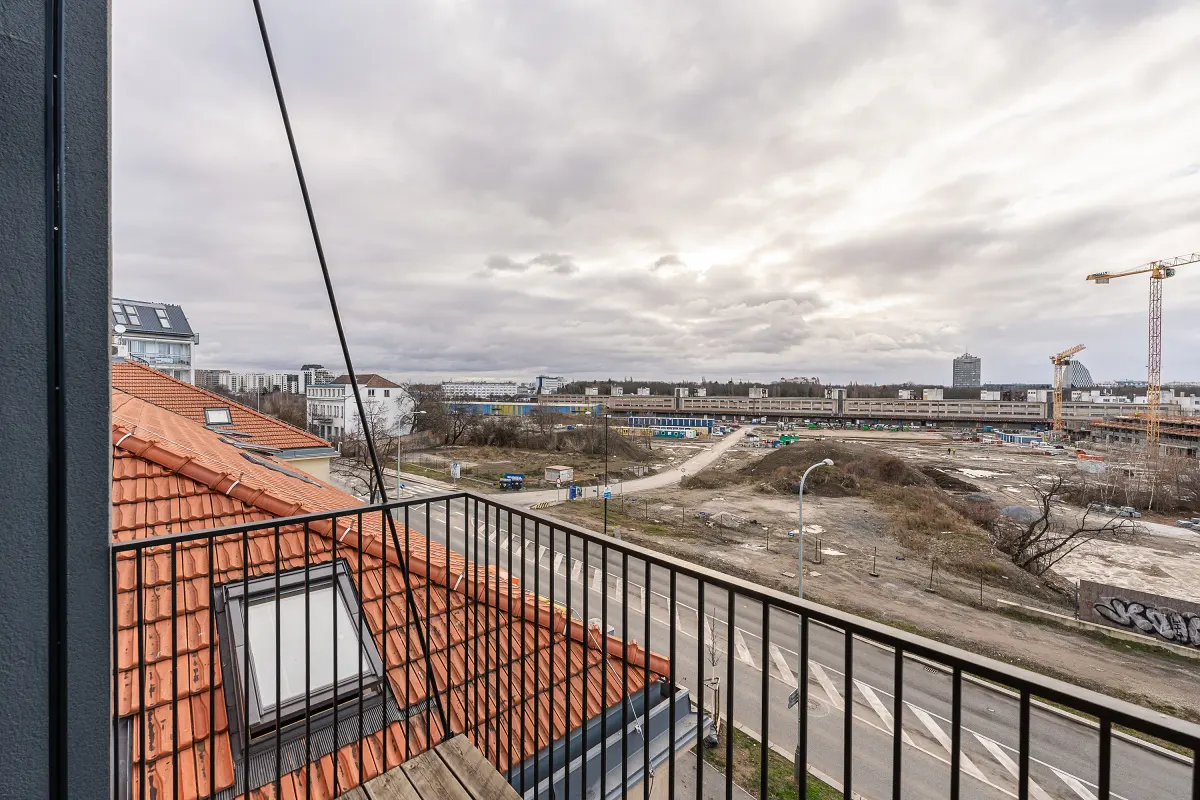 View from a balcony with black railings, overlooking a red tile roof, a road, and a construction site with cranes under a cloudy sky.