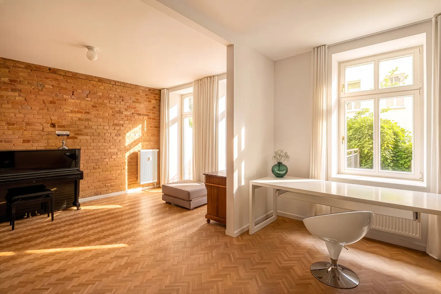 Bright, open-concept room with herringbone wood floors, brick wall, and piano. A white desk and chair sit by a window with sheer curtains.