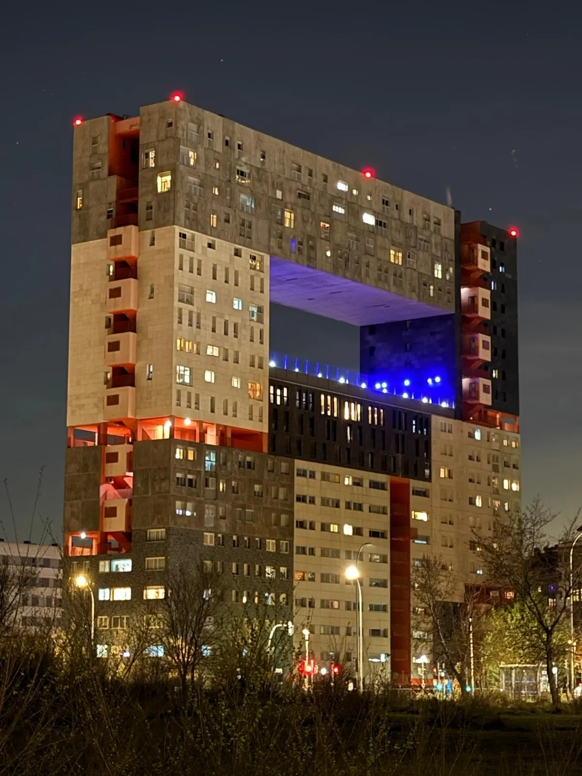 Night view of the Mirador Building in Madrid, a unique concrete structure with a large central opening. Windows glow with warm light.