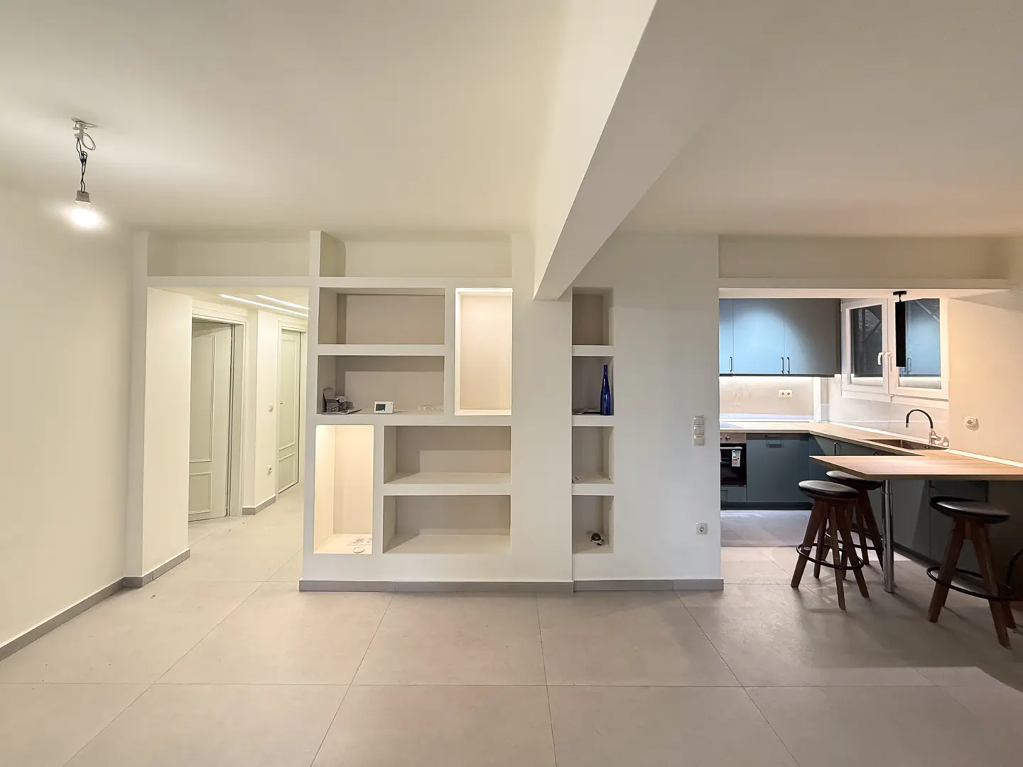 A bright, modern apartment interior with white walls, built-in shelving, and a kitchen with dark cabinets and wooden stools.