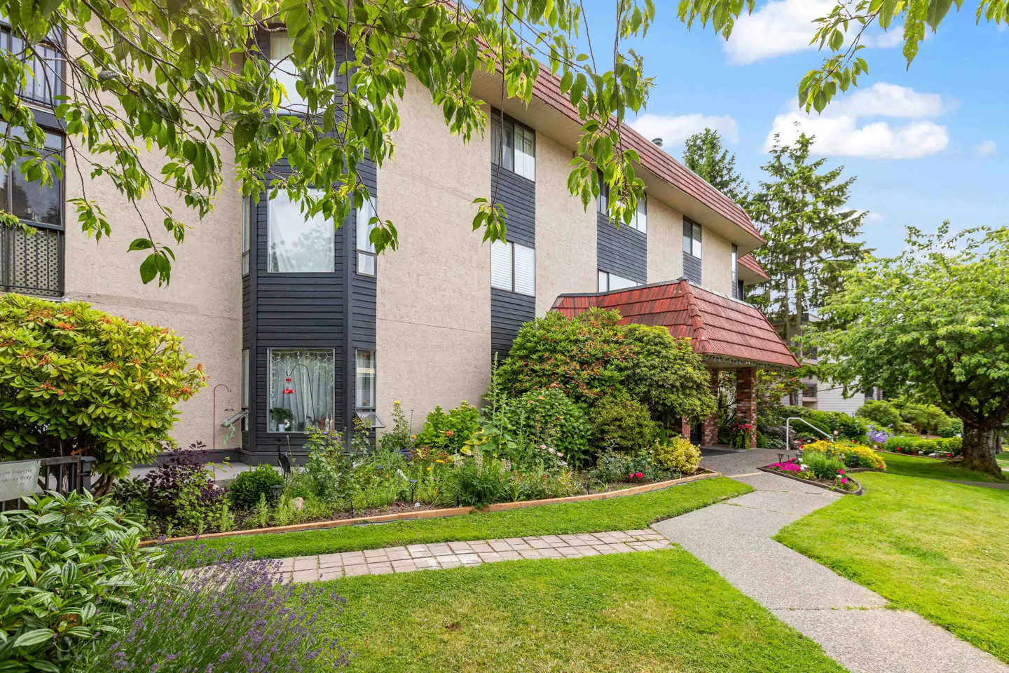 Exterior view of a beige and black apartment building with a red roof, surrounded by green grass, trees, and flowers.