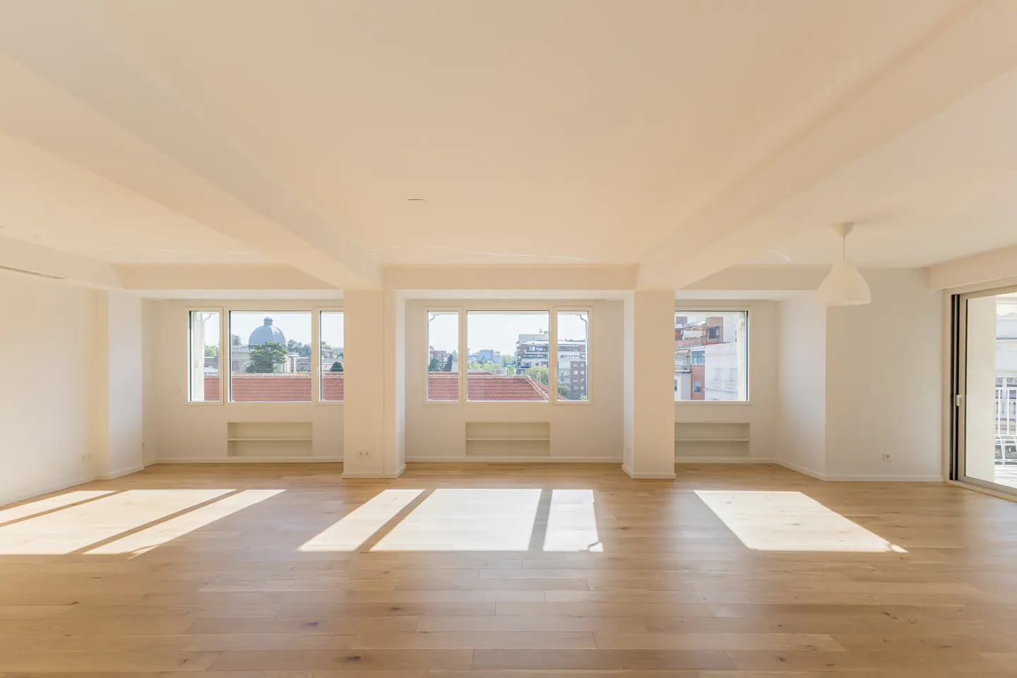 Bright, empty room with hardwood floors, white walls, and large windows showing a cityscape. Sunlight streams in, creating shadows on the floor.