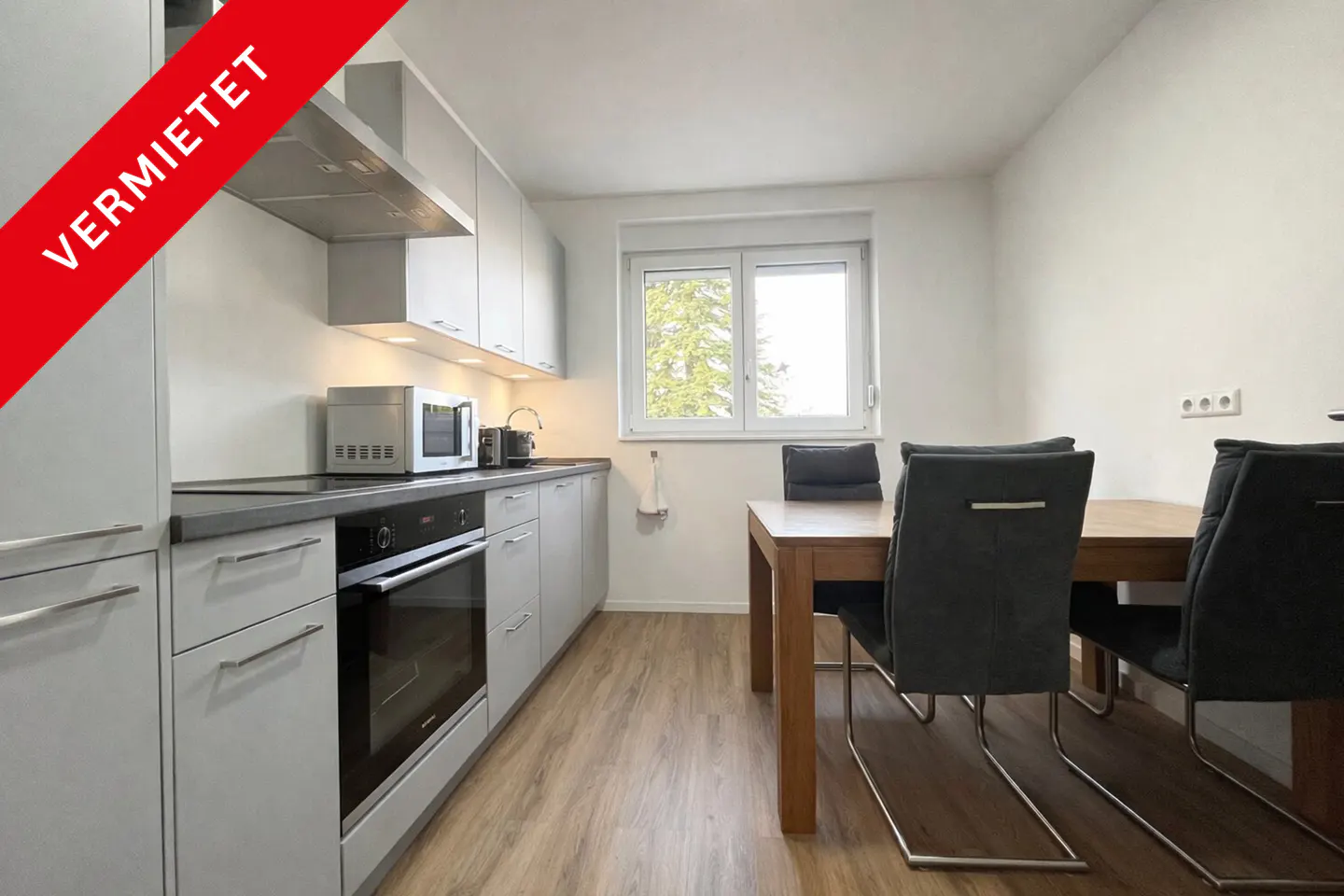 Bright kitchen with gray cabinets, stainless steel appliances, and a wooden dining table with four gray chairs. "VERMIETET" banner in the upper left corner.