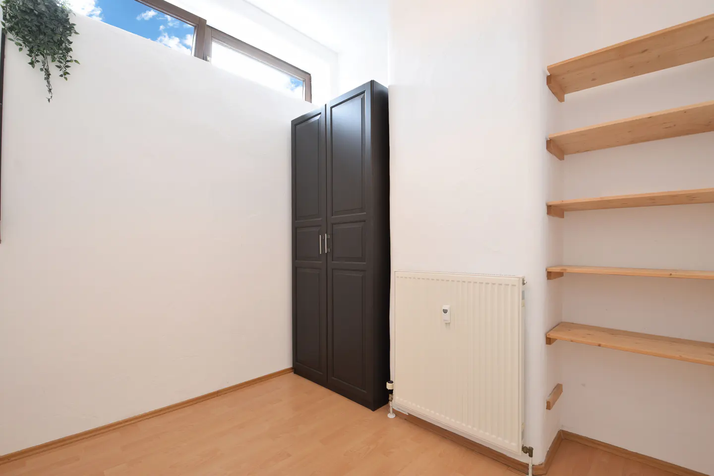 A room with light wood floors, white walls, a dark brown wardrobe, a white radiator, and wooden shelves.