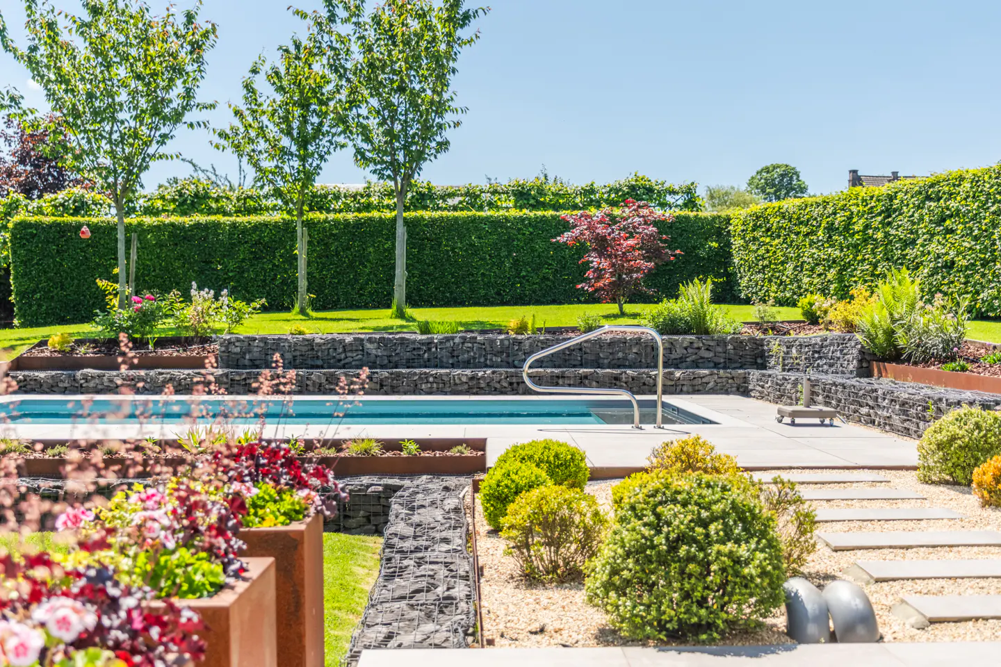A backyard with a pool, stone walls, and lush greenery under a clear blue sky.