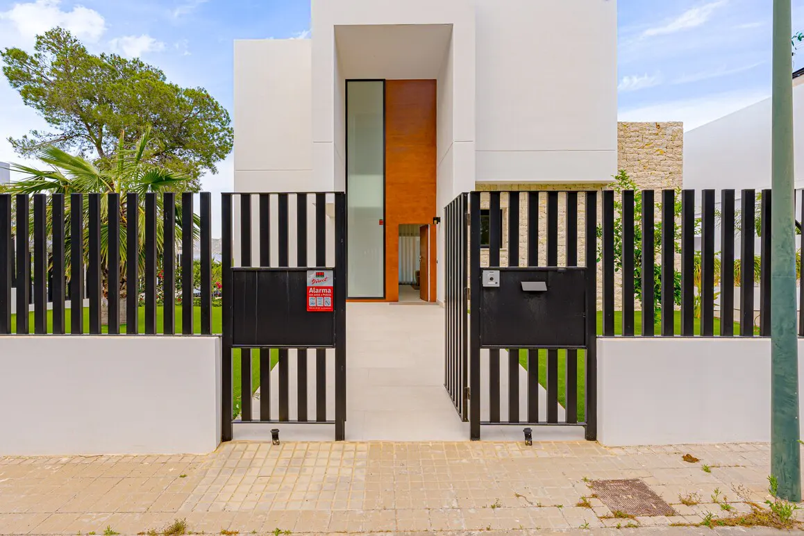 Open black metal gate leads to a modern white house with a tall wood-paneled entryway. Green lawn and trees visible behind the gate.
