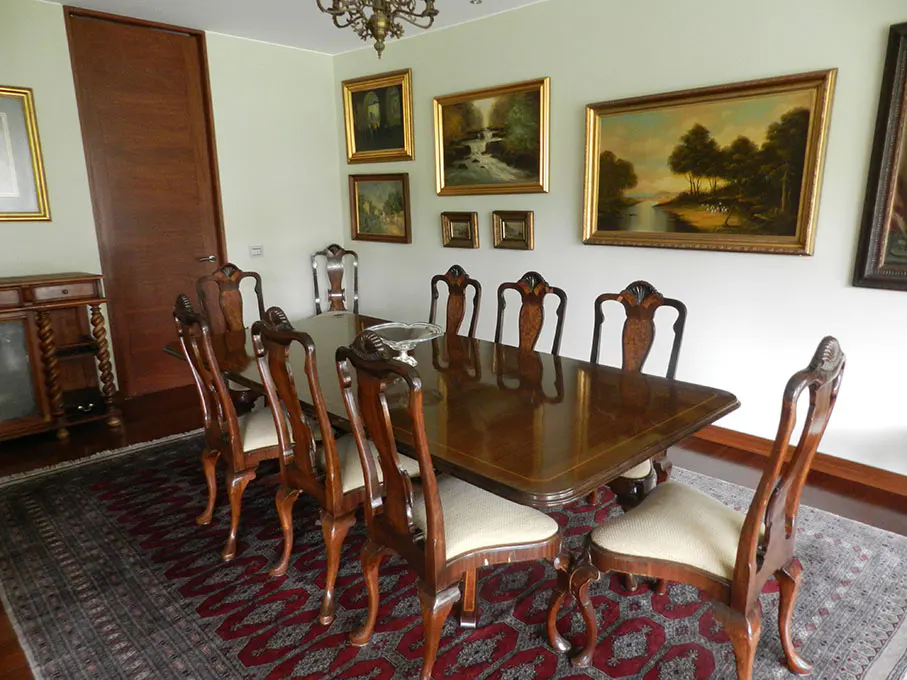 A formal dining room with a long, dark wood table surrounded by ornate chairs. Paintings adorn the light green walls. A patterned rug covers the floor.