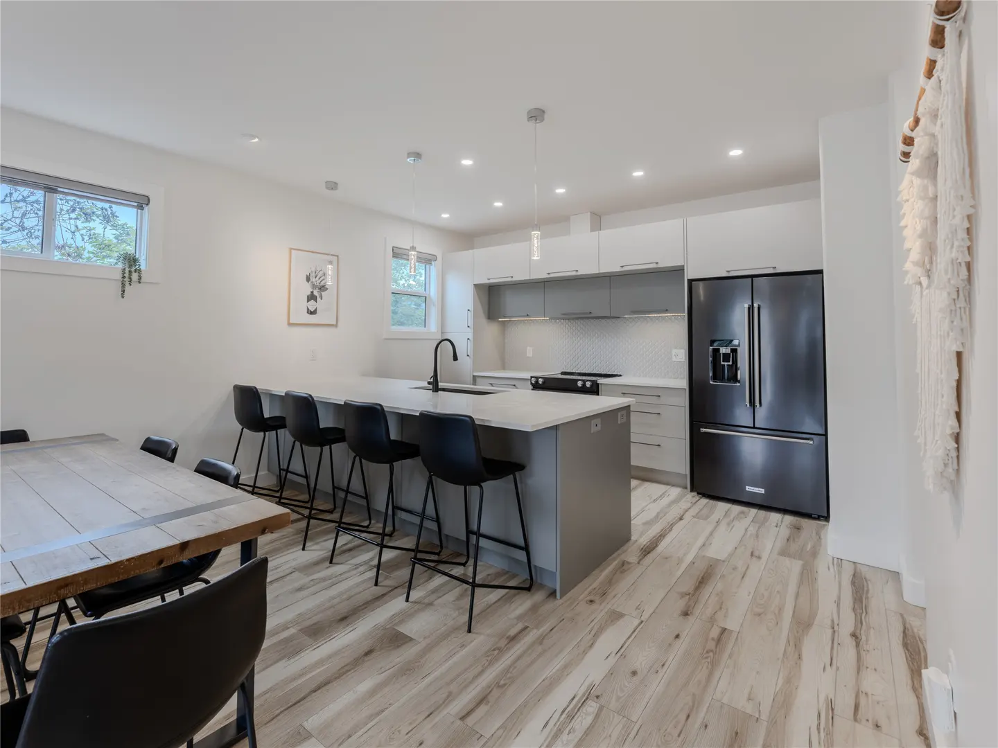 Modern kitchen with gray cabinets, white countertops, black bar stools, a black refrigerator, and a rustic dining table.