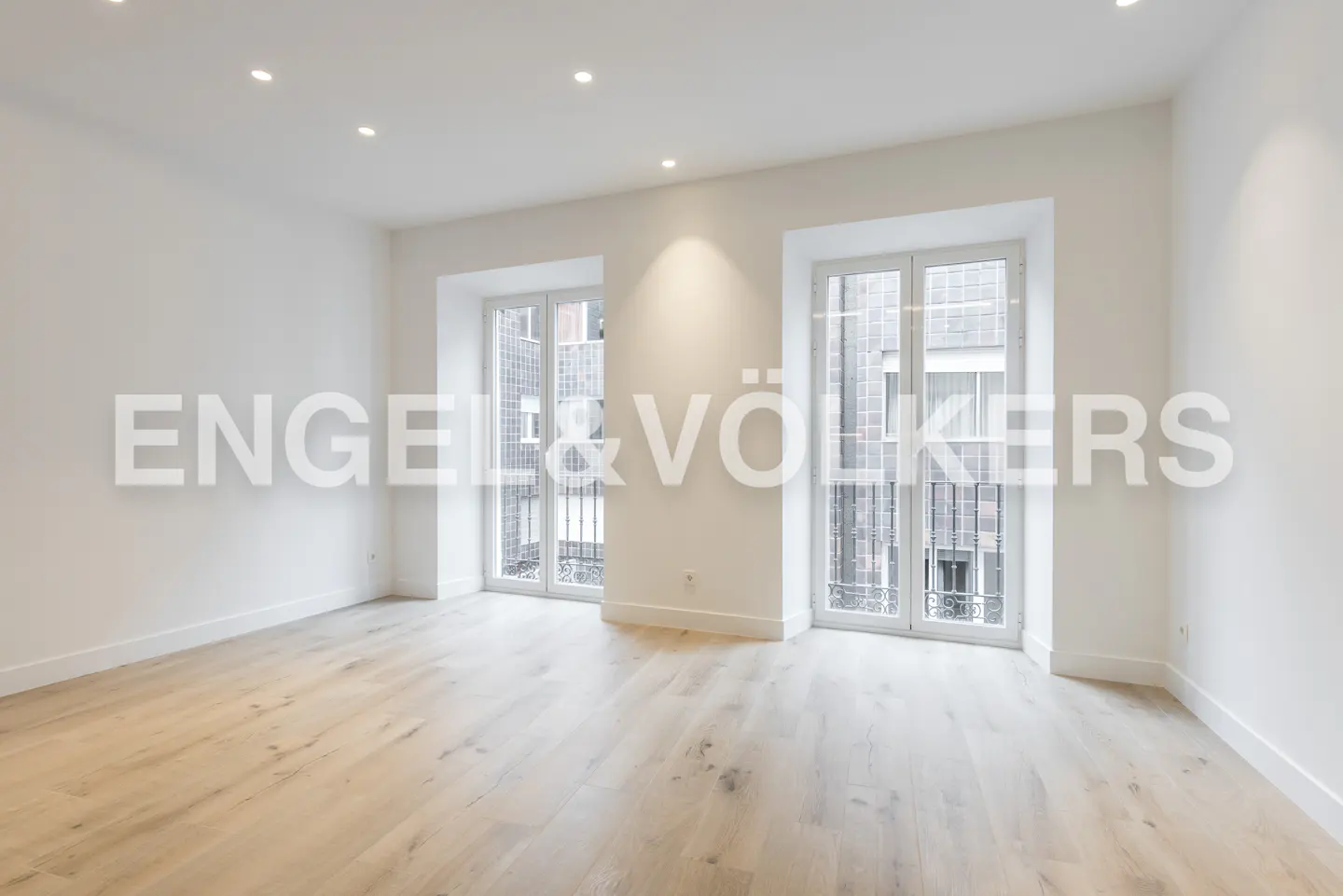 Bright, empty room with light wood floors, white walls, and two windows with black iron railings. Engel & Völkers logo overlay.