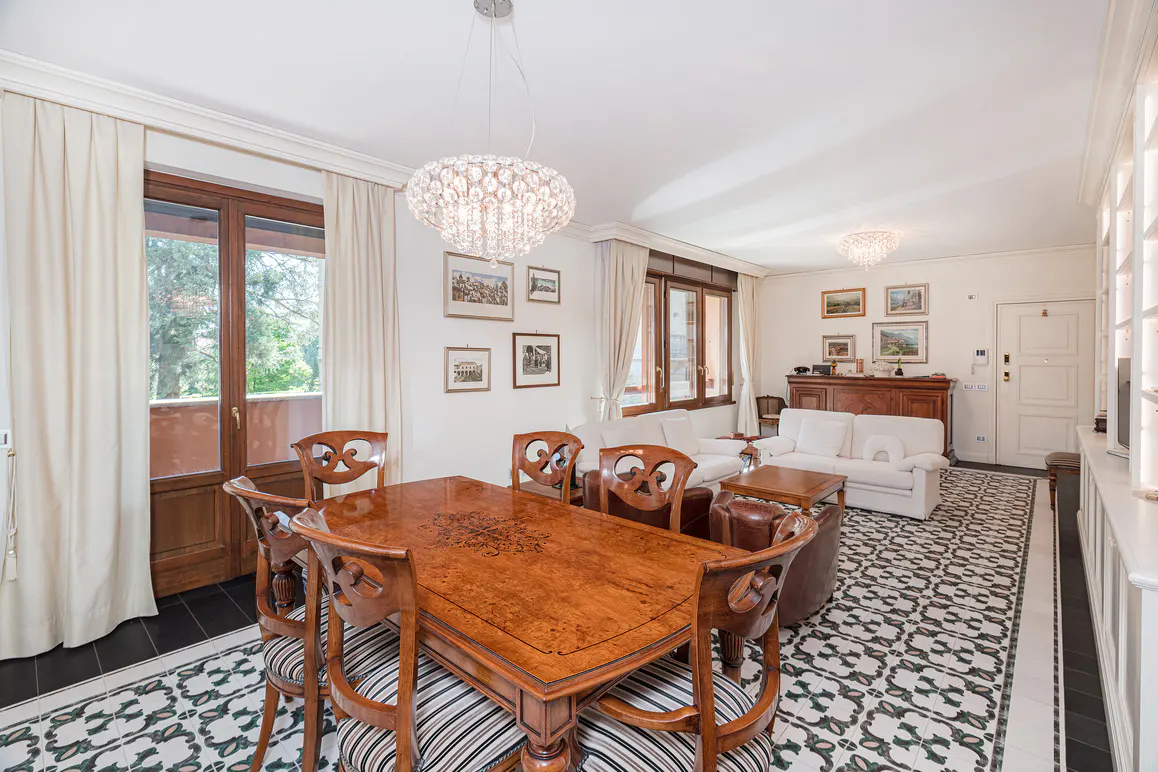 Elegant dining room with a polished wood table, patterned tile floors, crystal chandeliers, and white sofas in the back.