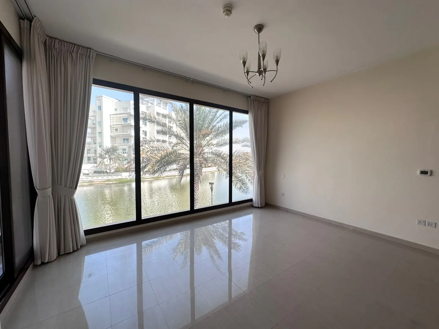 Empty room with tile floor, beige walls, and a large window overlooking a lake with palm trees and buildings. Chandelier on the ceiling.