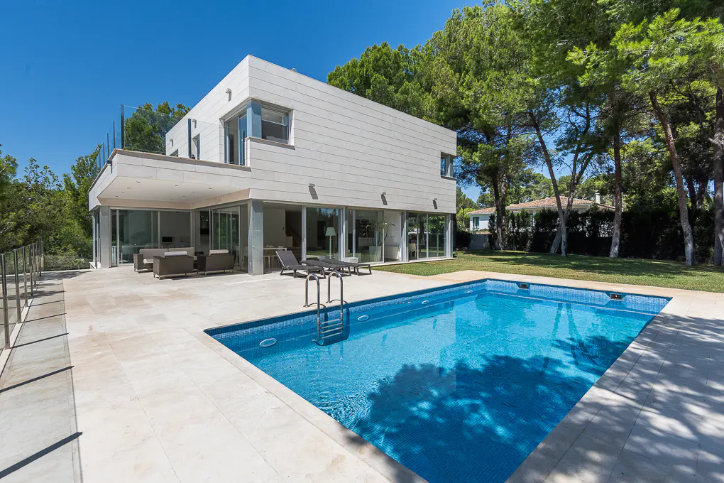 Modern white house with a blue tiled pool, patio furniture, and green trees under a clear blue sky.