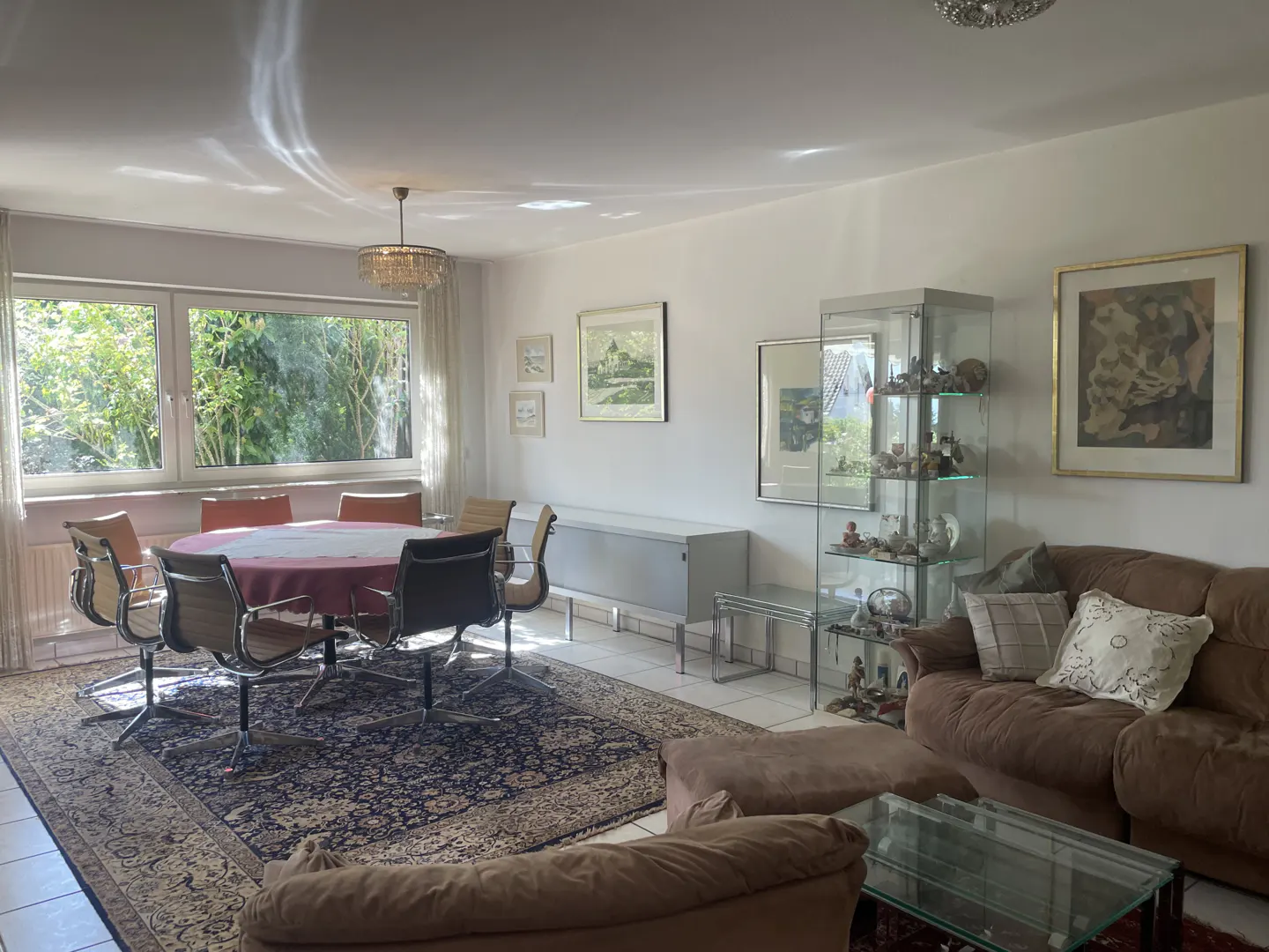 Bright living room with a round table, chairs, brown sofa, glass display case, and a patterned rug. Sunlight streams through a large window.