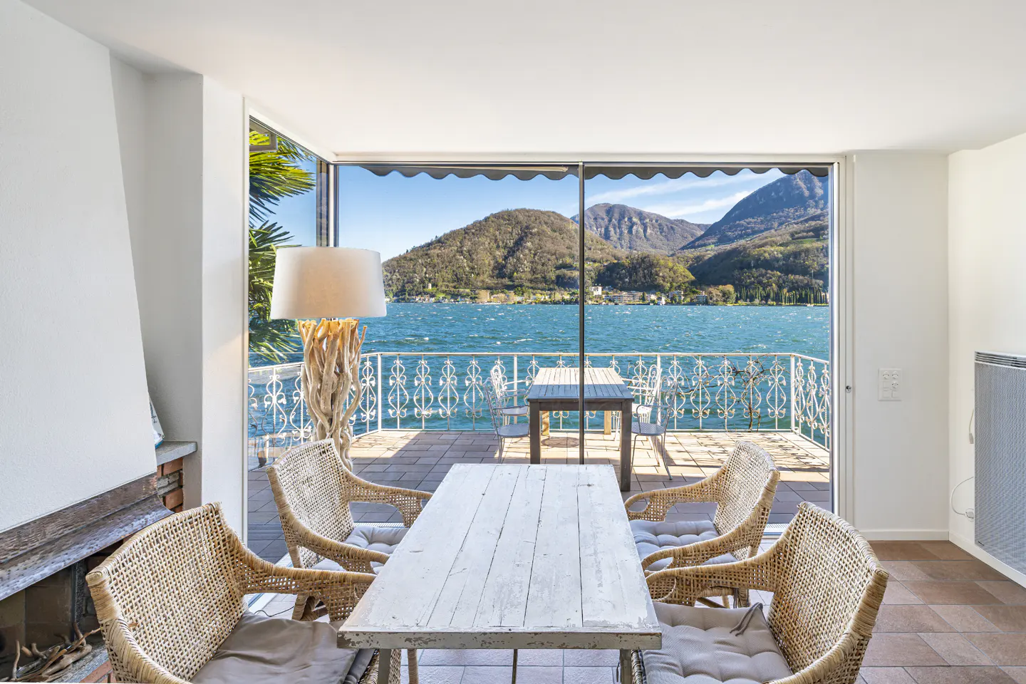 Dining area with white wooden table and wicker chairs, opening to a balcony overlooking a lake and mountains.