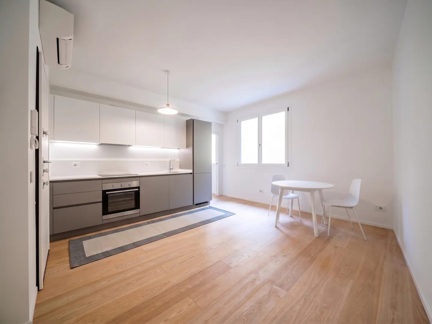 Bright, modern kitchen with white and gray cabinets, oven, and sink. A white table and chairs sit on wood floors near a window.