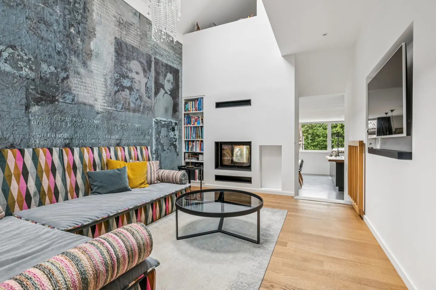 Modern living room with a patterned sofa, glass coffee table, fireplace, built-in bookshelf, and artistic mural wall.