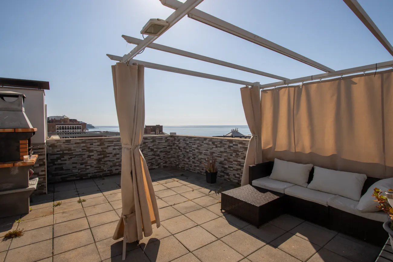 Rooftop patio with a pergola, beige curtains, and a dark brown wicker sofa with white cushions. A brick wall and ocean view are in the background.