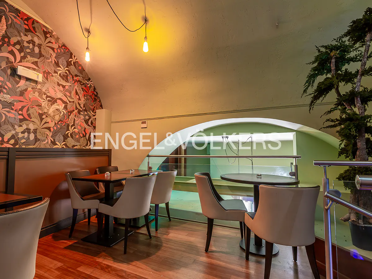 Interior view of a restaurant with tables, chairs, and floral wallpaper. A large plant is visible near a glass railing.