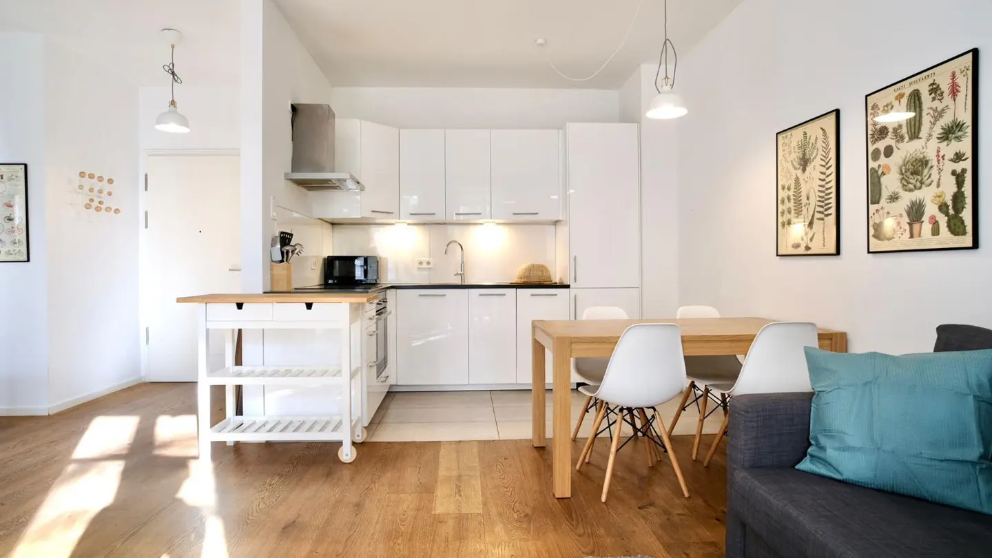 Bright, modern apartment interior with white kitchen, wood floors, dining table with white chairs, and a gray sofa with a blue pillow.
