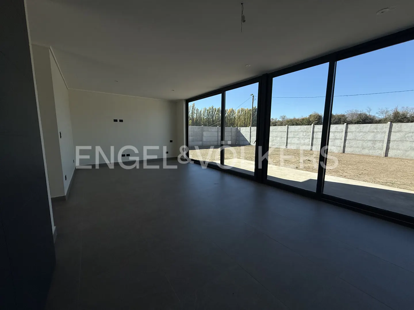 An empty room with gray floors and white walls. Large black-framed windows look out onto a yard with a concrete wall. "Engel & Volkers" logo is superimposed.