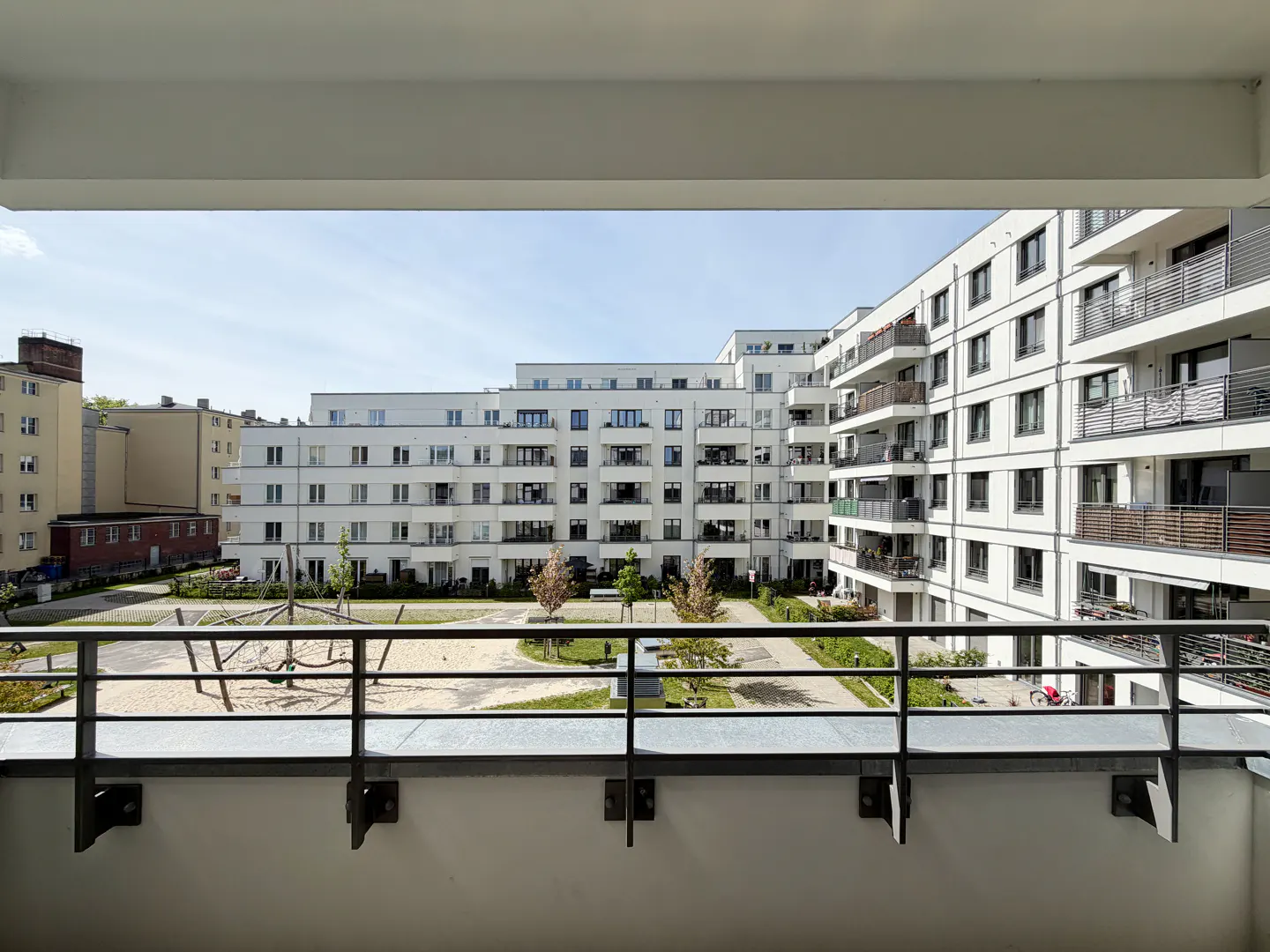 View from a balcony overlooking a courtyard with white apartment buildings and a playground on a sunny day.