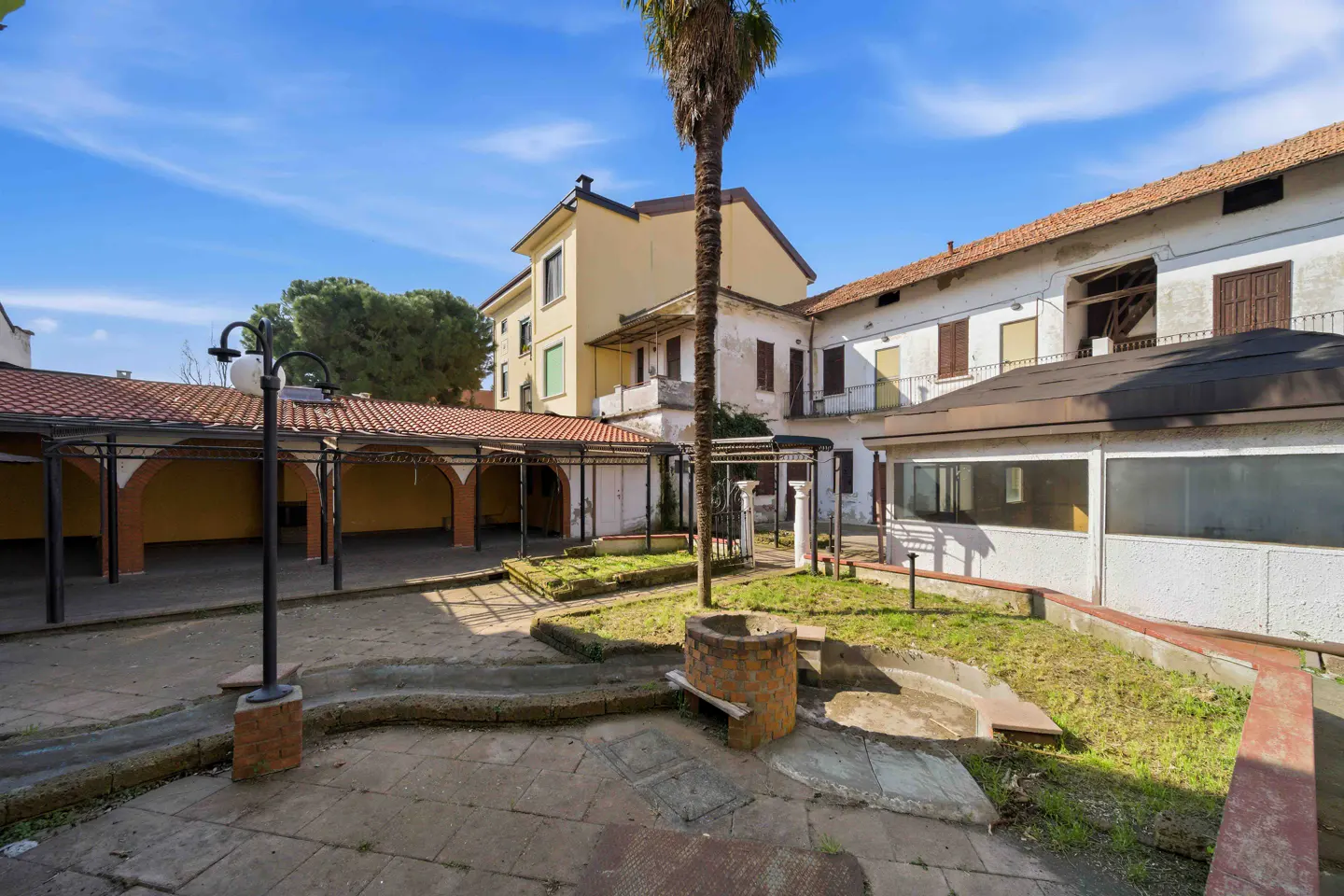 Exterior view of a courtyard with buildings, a palm tree, and a covered walkway on a sunny day.