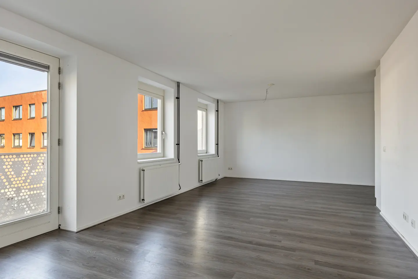 Bright, empty room with gray wood floors, white walls, and three windows. An orange building is visible through the windows.