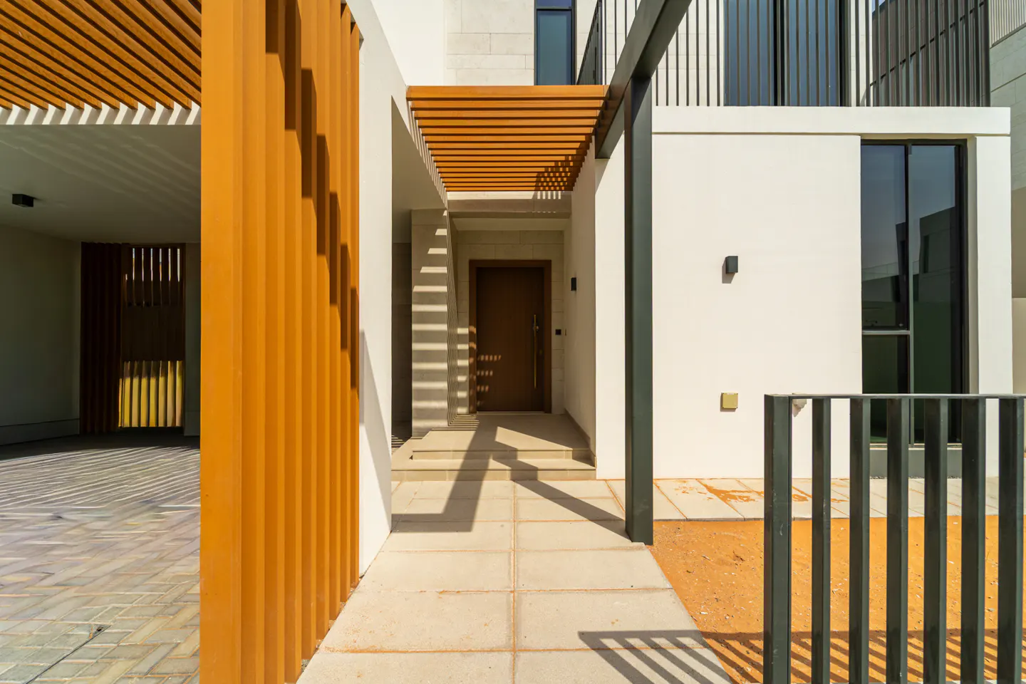 Modern home exterior with a brown front door, white walls, and wooden slat accents casting shadows on the walkway.
