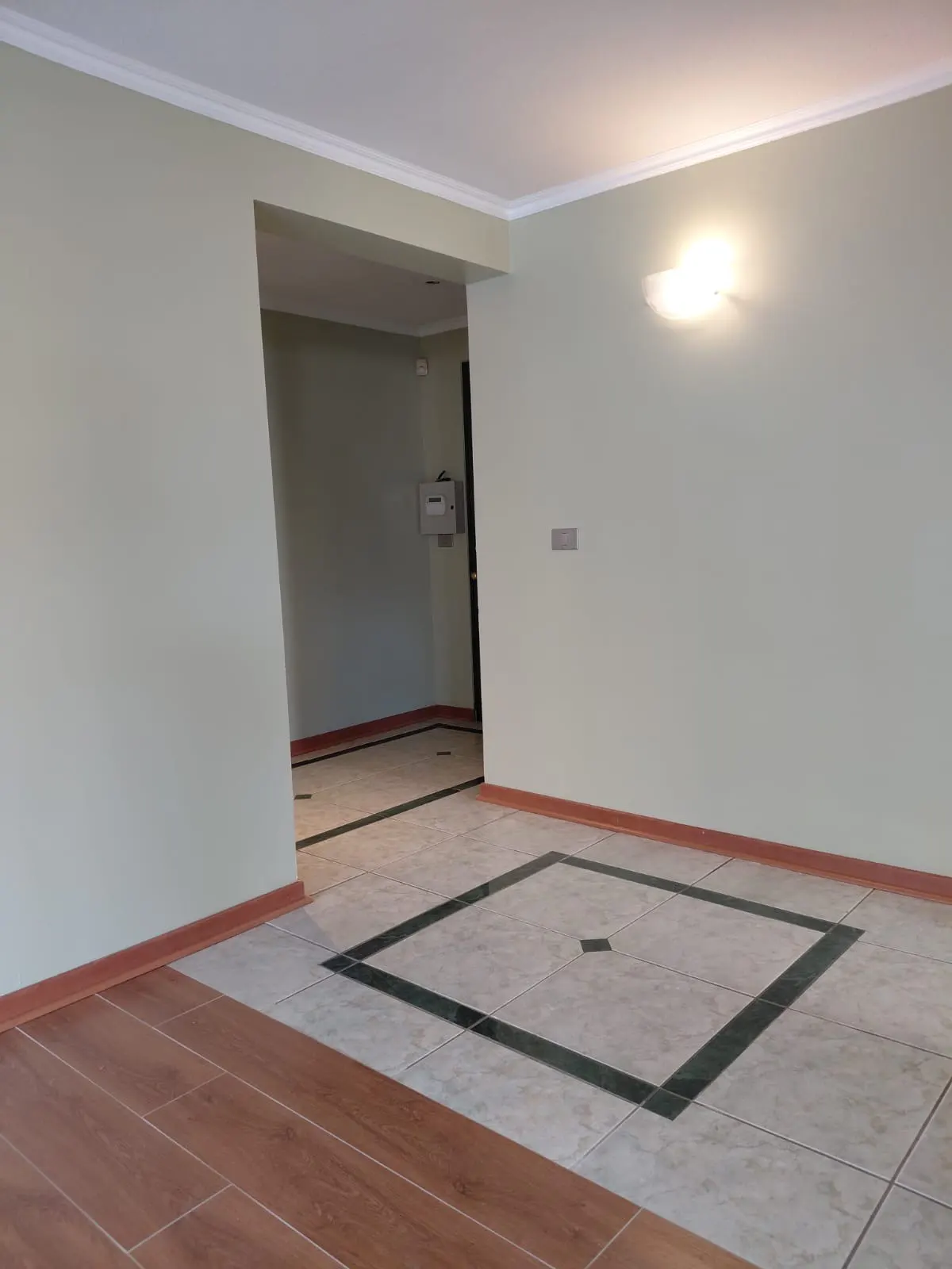 An empty room with light green walls, white trim, and tile flooring with a dark green square design. A doorway leads to another room.