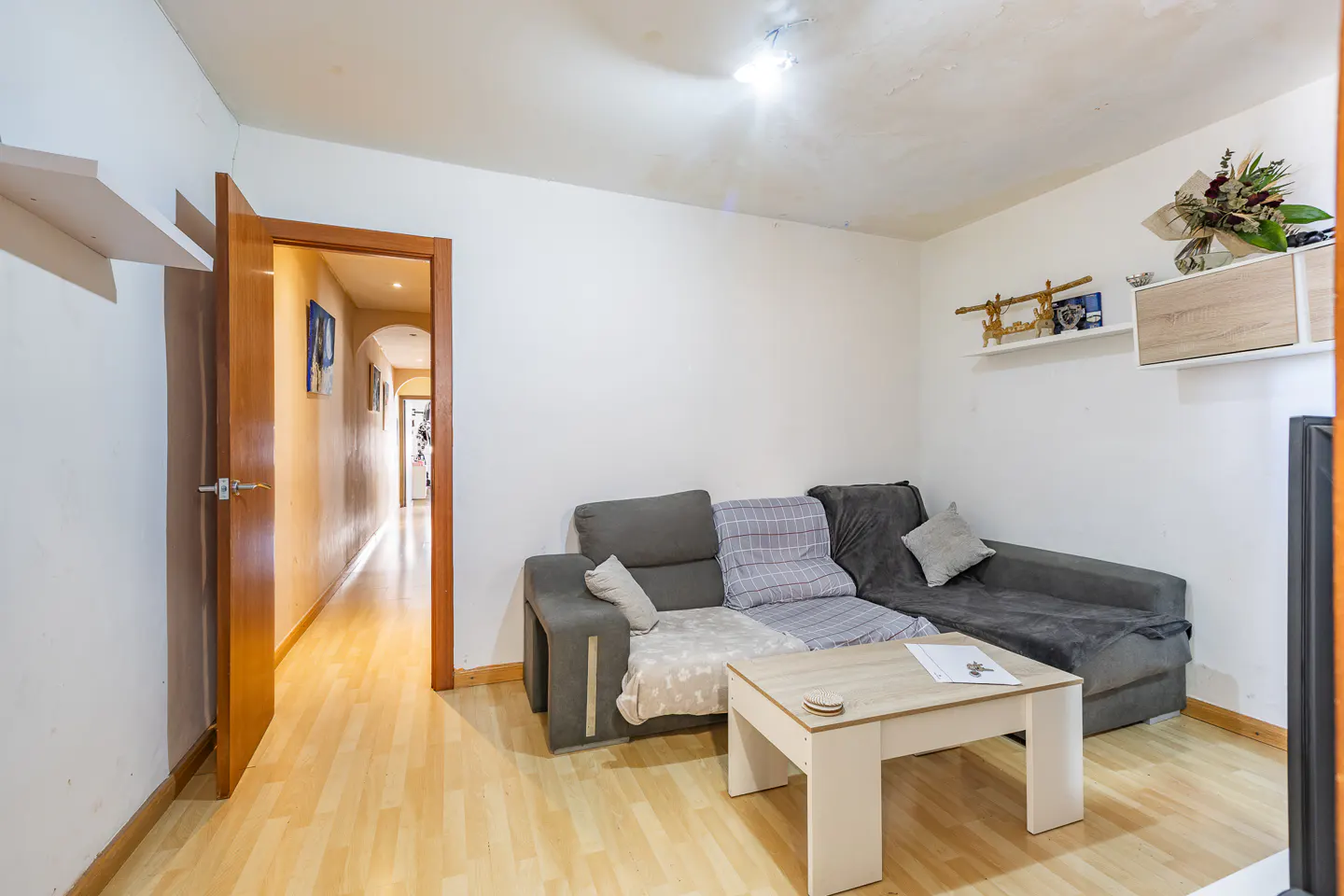 Living room with gray sectional sofa, white walls, and light wood floors. A white coffee table sits in front of the sofa. A hallway is visible through an open door.