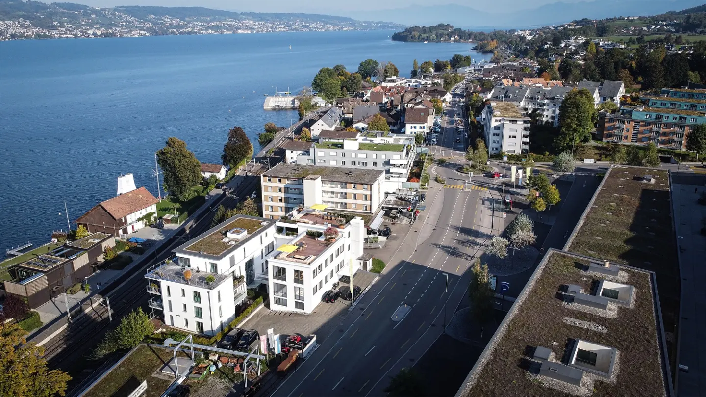 Aerial view of a lakeside town with modern white buildings, a road, and a large blue lake in the background.