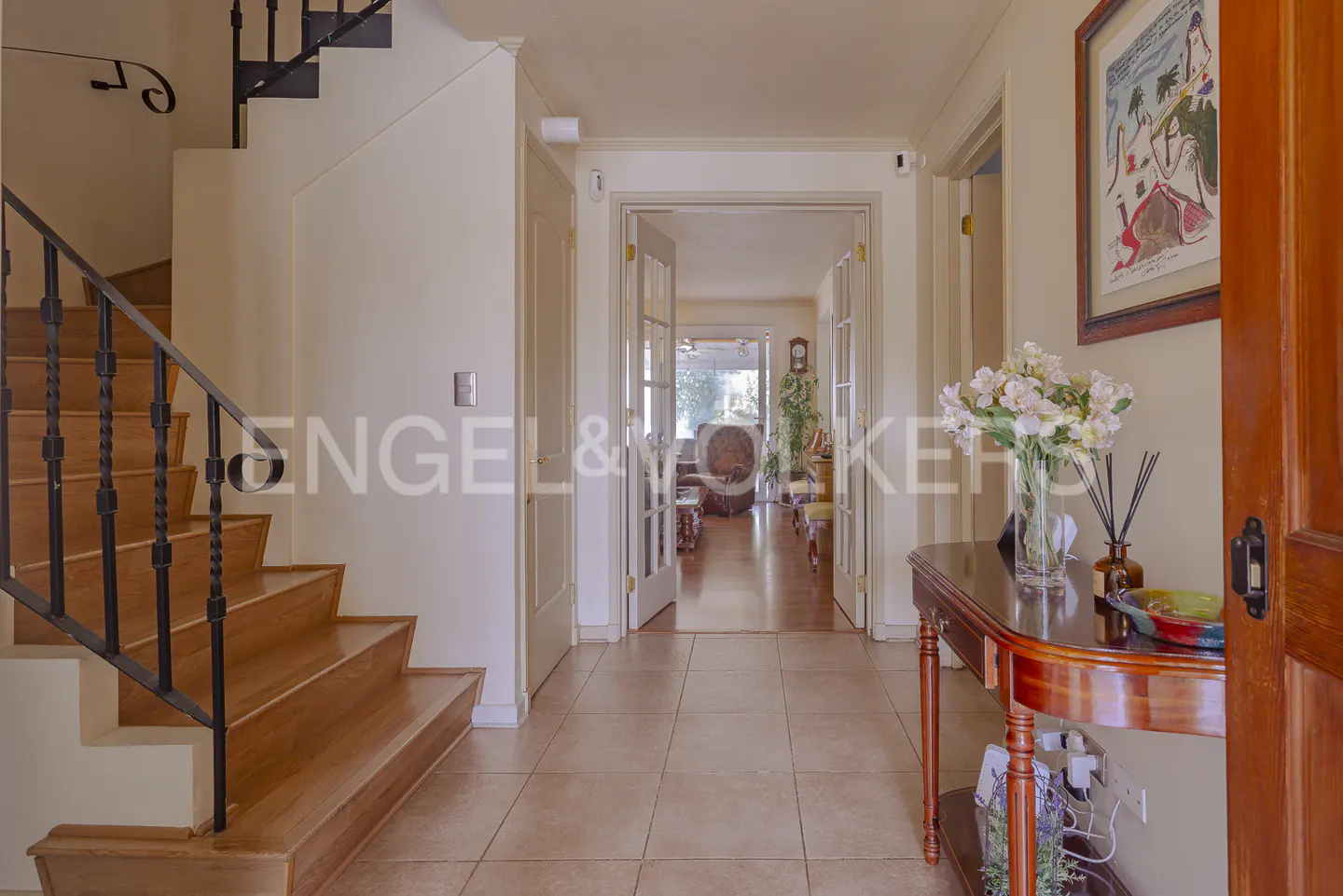 Entryway with wood stairs, tile floor, and a view through glass doors to a living room. A wood table with flowers sits near the front door.