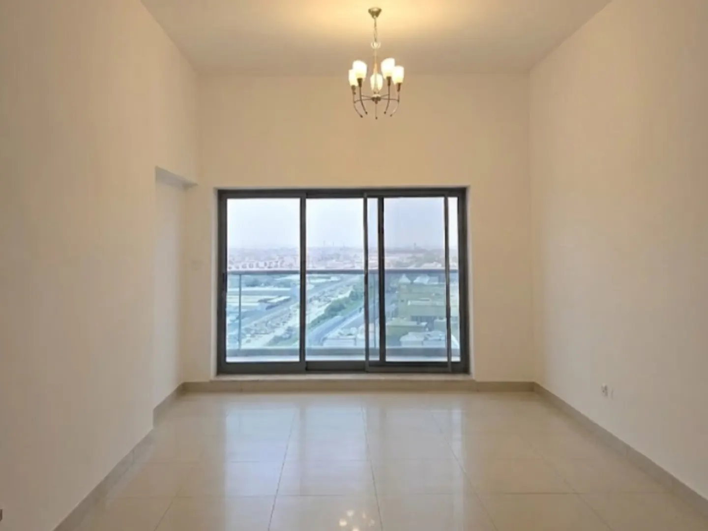 Empty room with beige walls and tile floor. A chandelier hangs from the ceiling. Large sliding glass doors lead to a balcony with a city view.