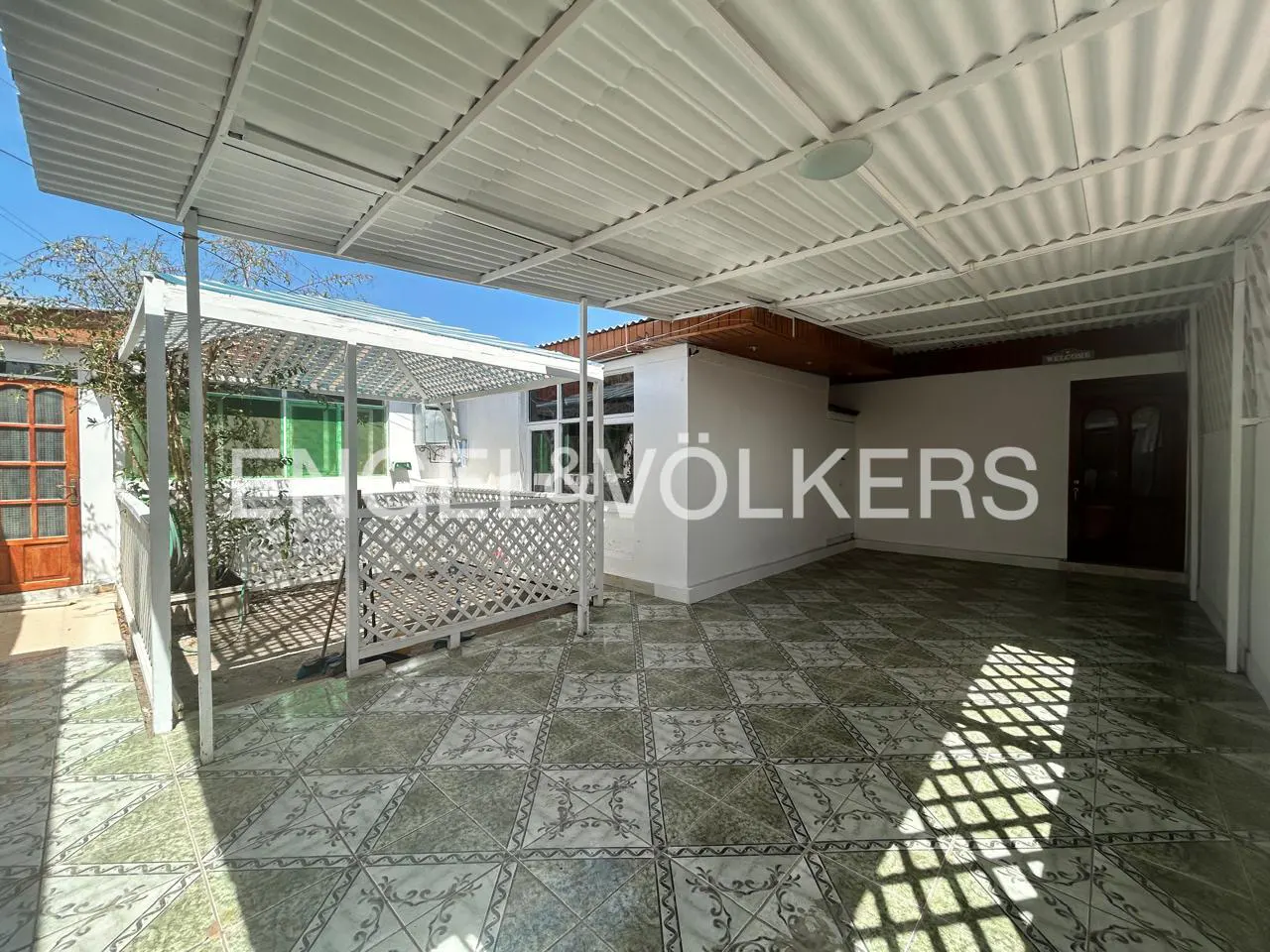 Covered patio with patterned tile flooring, white pergola, and white corrugated metal roof. A brown door is visible in the background.