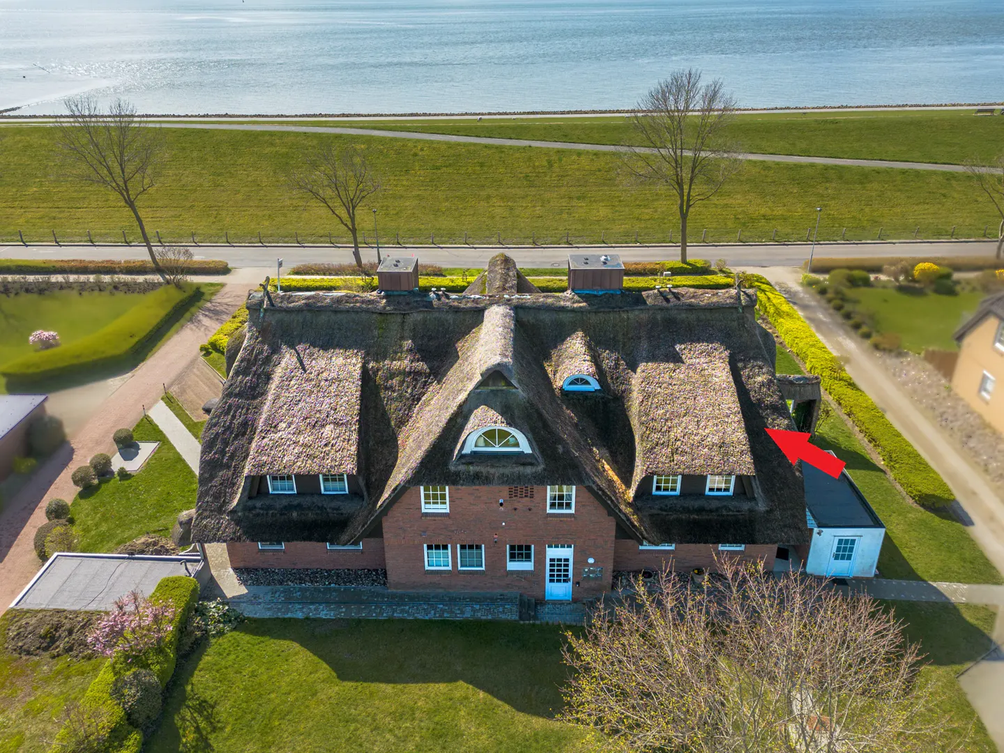 Aerial view of a brick house with a thatched roof, green lawn, and a body of water in the background.