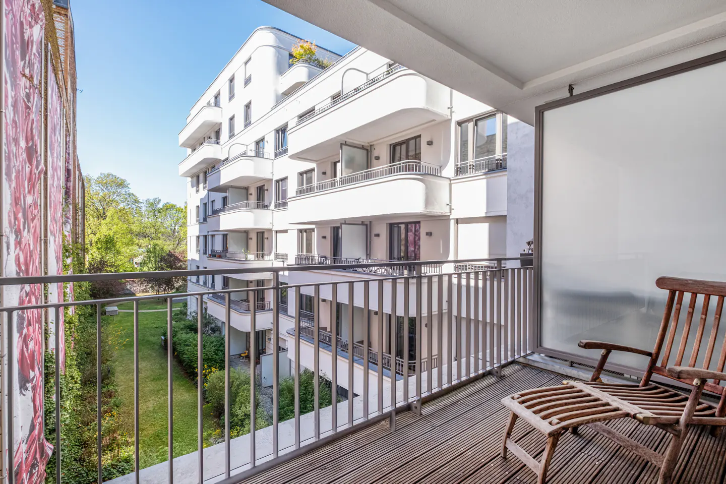 Balcony view with wooden deck, metal railing, and chair. White apartment building and green space visible in the background.