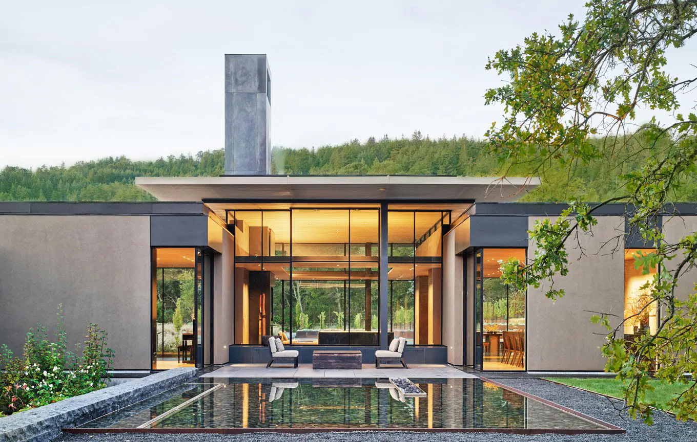 Modern home exterior with a reflecting pool, floor-to-ceiling windows, and a stone chimney against a green forest backdrop.