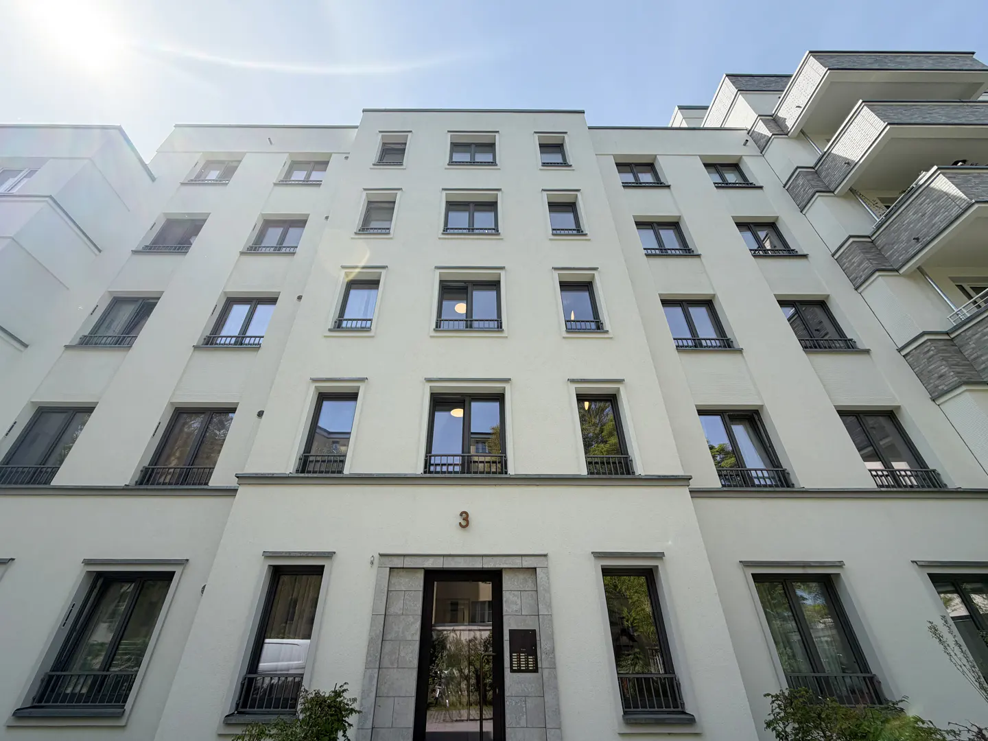 Low-angle shot of a white apartment building with black-framed windows under a blue sky. The number 3 is above the entrance.