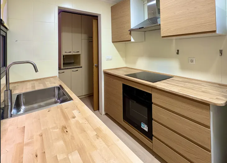 A bright kitchen with light wood cabinets, stainless steel sink, and black stovetop.