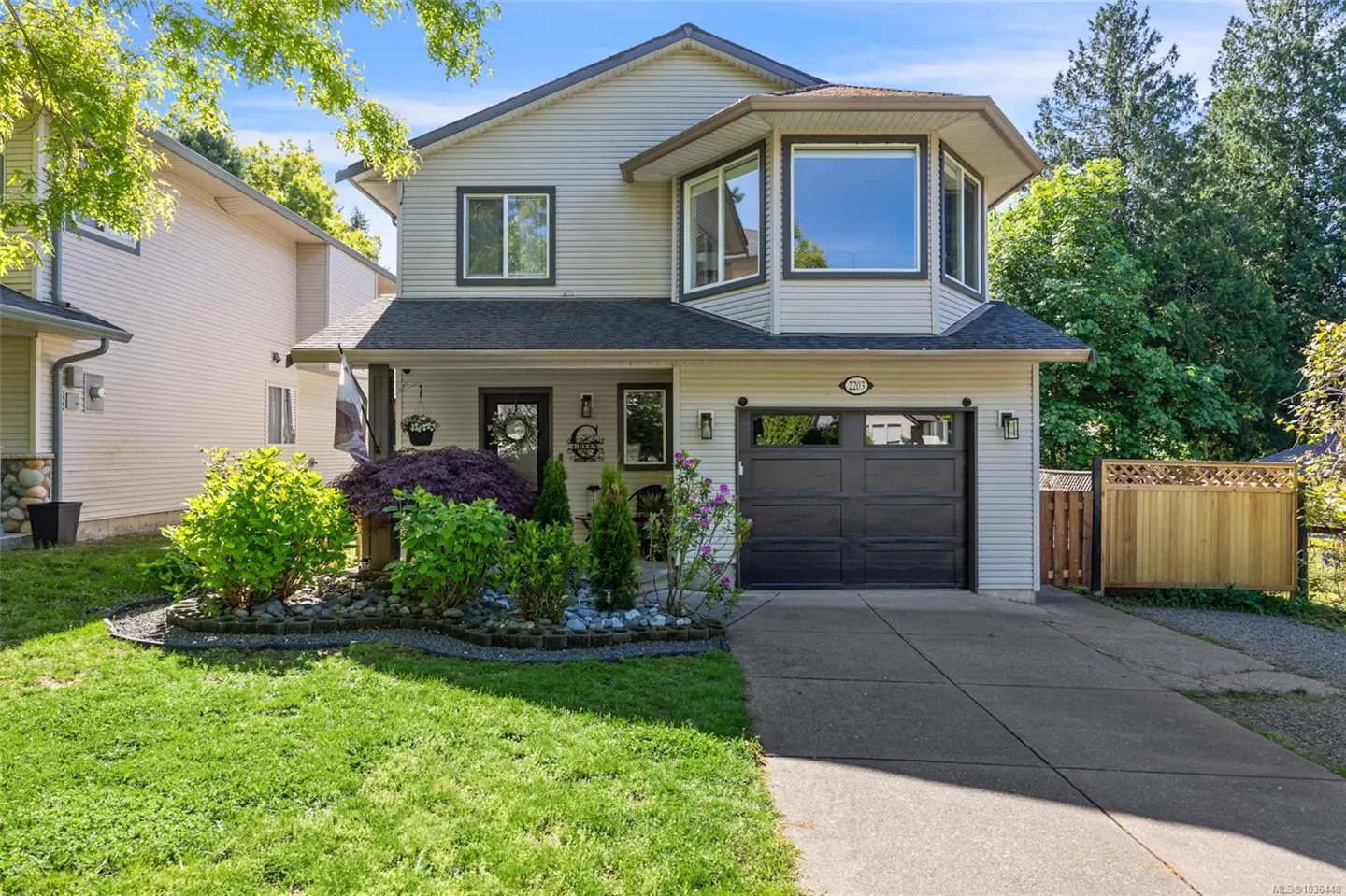 Two-story beige house with a dark gray garage door, green lawn, and landscaped garden.