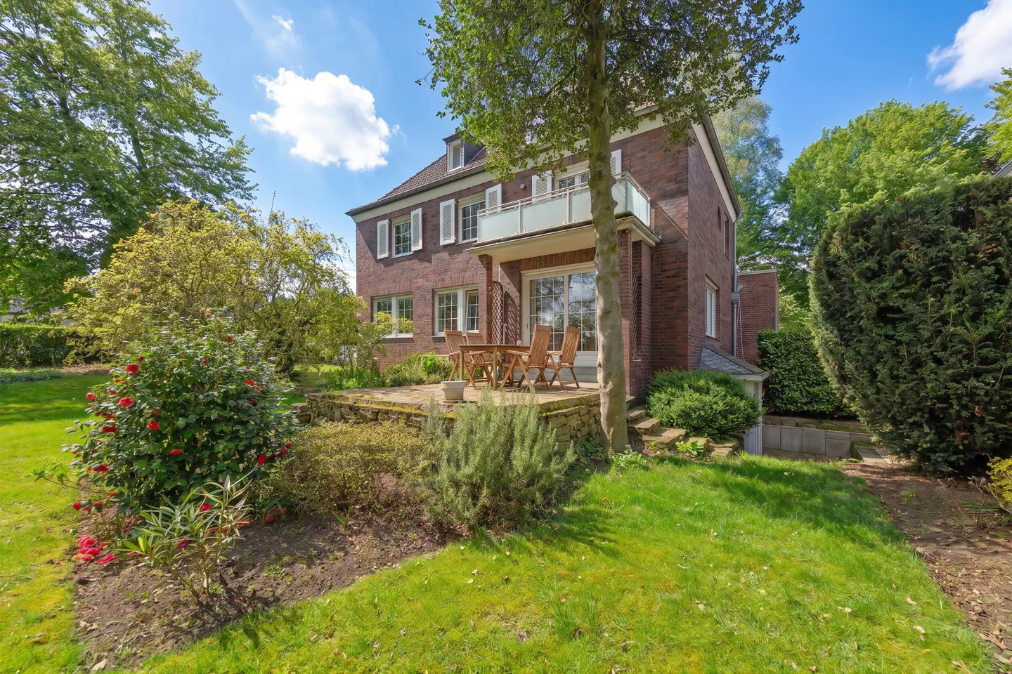Exterior view of a two-story red brick house with a balcony and patio with wooden furniture. Green lawn and trees surround the house.