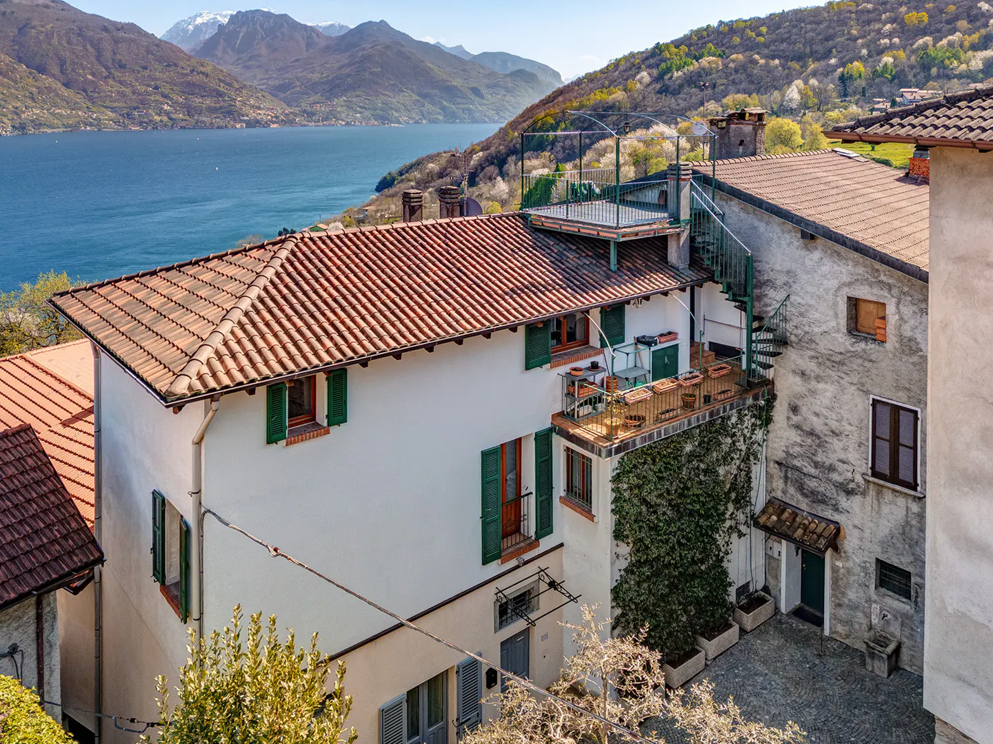 Scenic view of Italian houses with red tile roofs and green shutters, overlooking a blue lake and mountains.