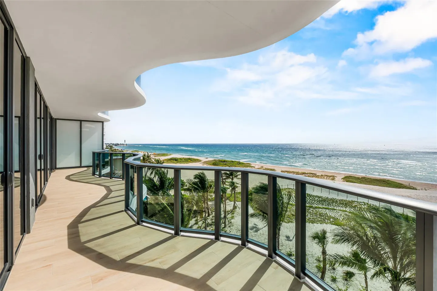 Curved balcony with glass railing overlooking a beach and ocean under a blue sky.