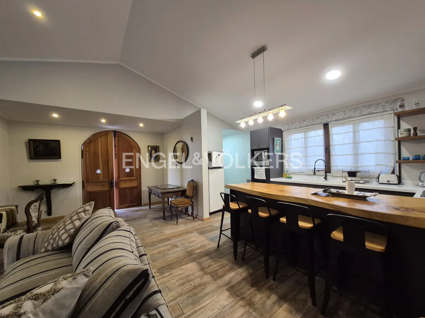 Open-concept living space with a striped couch, wood floors, and a kitchen island with black stools. Arched wooden doors are visible.