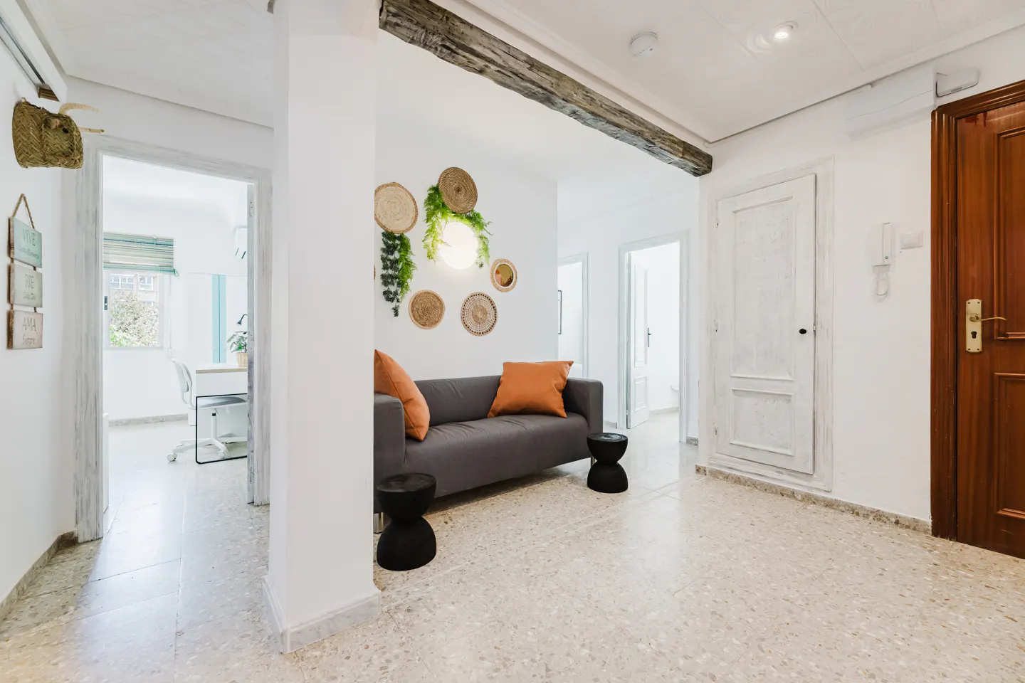 Bright hallway with a gray sofa, orange pillows, and woven wall decor. A wooden beam accents the white walls and terrazzo floor.